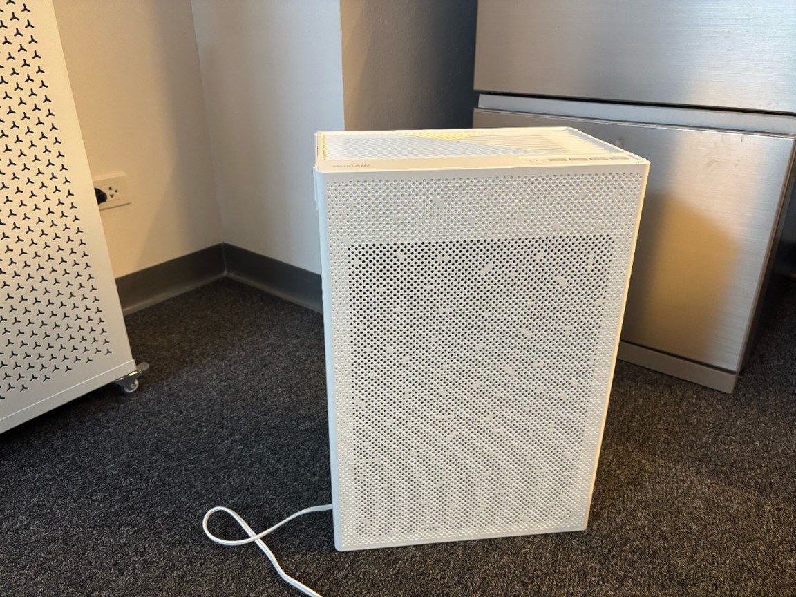 Smart Air SA600 ตั้งบนพรมสีเข้มในมุมออฟฟิศ มีสายไฟลากอยู่ ข้างตู้เหล็กและด้านข้างของ Smart Air Blast