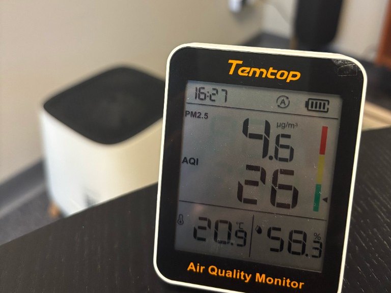 เครื่องวัดคุณภาพอากาศ Temtop S1 บนโต๊ะในห้องทดสอบ แสดงค่า PM2.5 4.6 µg/m³ มี Smart Air Sqair วางบนพื้นด้านหลัง
