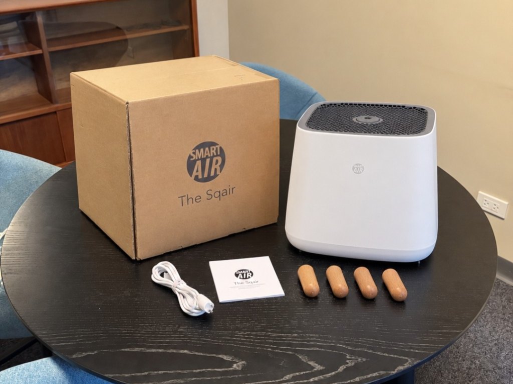 แกะกล่อง Sqair บนโต๊ะไม้กลมสีเข้ม มีตัวเครื่องสีขาว กล่องกระดาษ Smart Air ขาไม้สี่ขา สายไฟ และคู่มือ