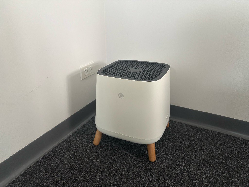 ภาพถ่ายมือถือ Smart Air Sqair บนขาไม้ในมุมห้องเล็กที่ปูพรม ข้างปลั๊กผนัง